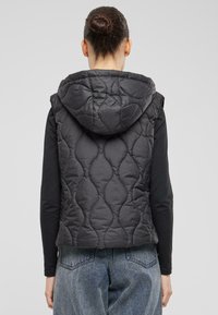 Schwarze gesteppte Weste mit Kapuze, die ein strukturiertes Muster aufweist, ärmelloses Design und eine taillierte Silhouette hat, kombiniert mit einem schwarzen Langarmshirt.