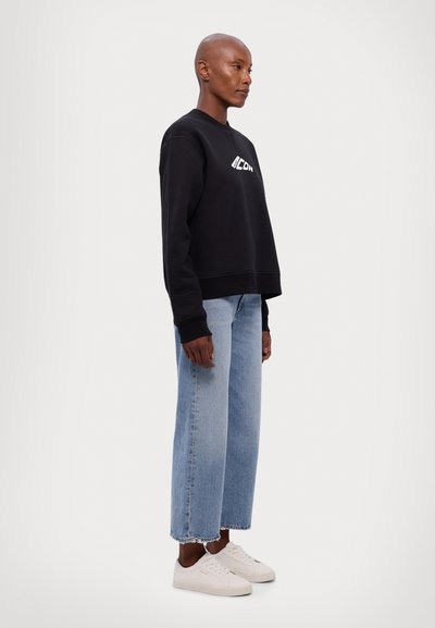 Black crew-neck sweatshirt with white logo, paired with light blue wide-leg jeans and white sneakers, showing a relaxed fit and cotton texture.