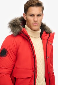 Chaqueta acolchada roja con capucha forrada de piel, cierre de cremallera en la parte delantera, acentos en negro y un parche en la manga, llevada sobre un suéter de punto con trenzas en color crema.