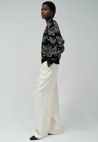 Pull noir avec broderie florale blanche, associé à un pantalon large blanc et des chaussures noires pointues. Le mannequin pose de côté devant un fond neutre.