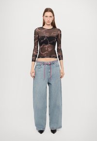 Schwarzes, durchsichtiges Mesh-Oberteil mit pinkem Blumenmuster, kombiniert mit hellblauen weiten Denim-Jeans, die einen pinken Kordelzug und Fronttaschen haben.