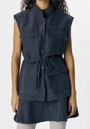 Mouwloze marineblauwe gilet met knopen, vier klepzakken en een tailleband, gedragen over een bijpassende rok met lichtgekleurde geribbelde panty's.