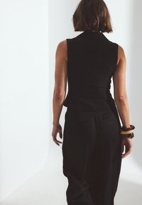 Must rääkimata top, millel on käteta lõige, kombineeritud lõdva musta pükste paariga. Aksessuaaride hulka kuuluvad värvilised käevõrud. Minimalistlik disain heledal taustal.