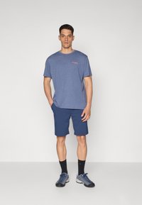 Blue short-sleeve T-shirt with small red logo, paired with navy shorts. Model wears gray sporty shoes with dark socks. Minimalist design.