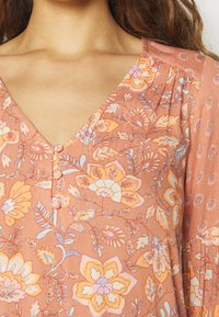 Kvinde iført en ferskenfarvet V-hals bluse med knapper og et blomsterdesign i orange, lilla og hvide nuancer.