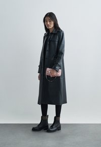 Manteau en cuir noir avec boutons, manches longues et col. Le modèle tient un petit sac à main rose. Leggings noirs et bottes de combat à lacets.