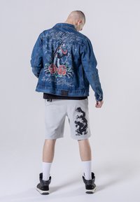 Denim jas met een geborduurde zwarte panter en rode rozen op de rug; grijze shorts met een zwart draakontwerp; hoge sneakers.