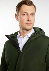Vihreä hupullinen takki, jossa on vetoketju ja napin sulkeminen, pehmeällä pinnalla ja mustalla reunuksella, käytetty valkoisen paidan päällä.