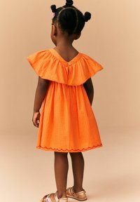 Oranje katoenen jurk met een gerimpelde halslijn en een geschulpte zoom. Heeft een knoopsluiting aan de achterkant en is gecombineerd met metalen sandalen.