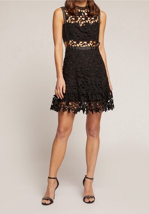 Femme en robe noire sans manches en dentelle avec des motifs floraux et des panneaux transparents, portant des sandales noires à talons hauts avec des brides à la cheville.