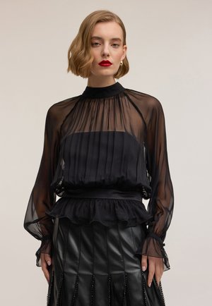 Donna con capelli corti biondi che indossa una blusa nera trasparente a maniche lunghe con orlo arricciato e una gonna plissettata in pelle nera.