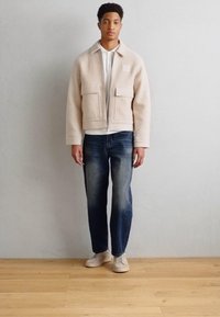 Veste en polaire couleur crème avec deux poches avant, associée à une chemise blanche et un jean bleu. Le modèle se tient sur un sol en bois.