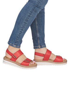 Osoba v modrých džínách a červených sandálech na otevřenou špičku s nastavitelnými pásky a bílými platformovými podrážkami, stojící s jednou nohou mírně zvednutou.