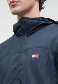 Κοντινό πλάνο ενός ατόμου που φοράει ένα ναυτικό μπλε ζακέτα με φερμουάρ και το λογότυπο Tommy Jeans στο στήθος, διαθέτοντας ψηλό γιακά και υφή υφάσματος.