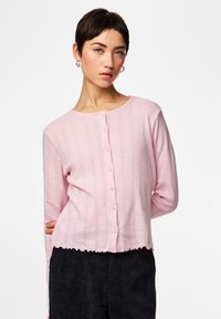 Femeie cu păr scurt și închis la culoare, purtând un cardigan roz deschis cu nasturi și pantaloni negri din catifea, stând în picioare pe un fundal alb simplu.