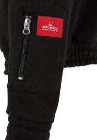 Sweatshirt noir avec des poignets élastiques, présentant une poche zippée argentée et une étiquette rouge avec du texte blanc et un logo de couronne.