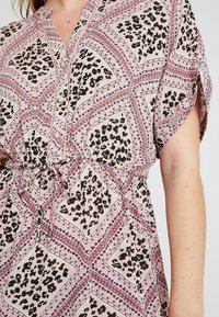 Woman wearing a pink patterned dress with black and purple geometric and leopard print designs, featuring a front tie and buttons.