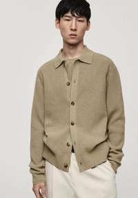 Cardigan lavorato a maglia beige con chiusura a bottoni, colletto aperto e maniche lunghe, abbinato a pantaloni crema, creando un look a strati.