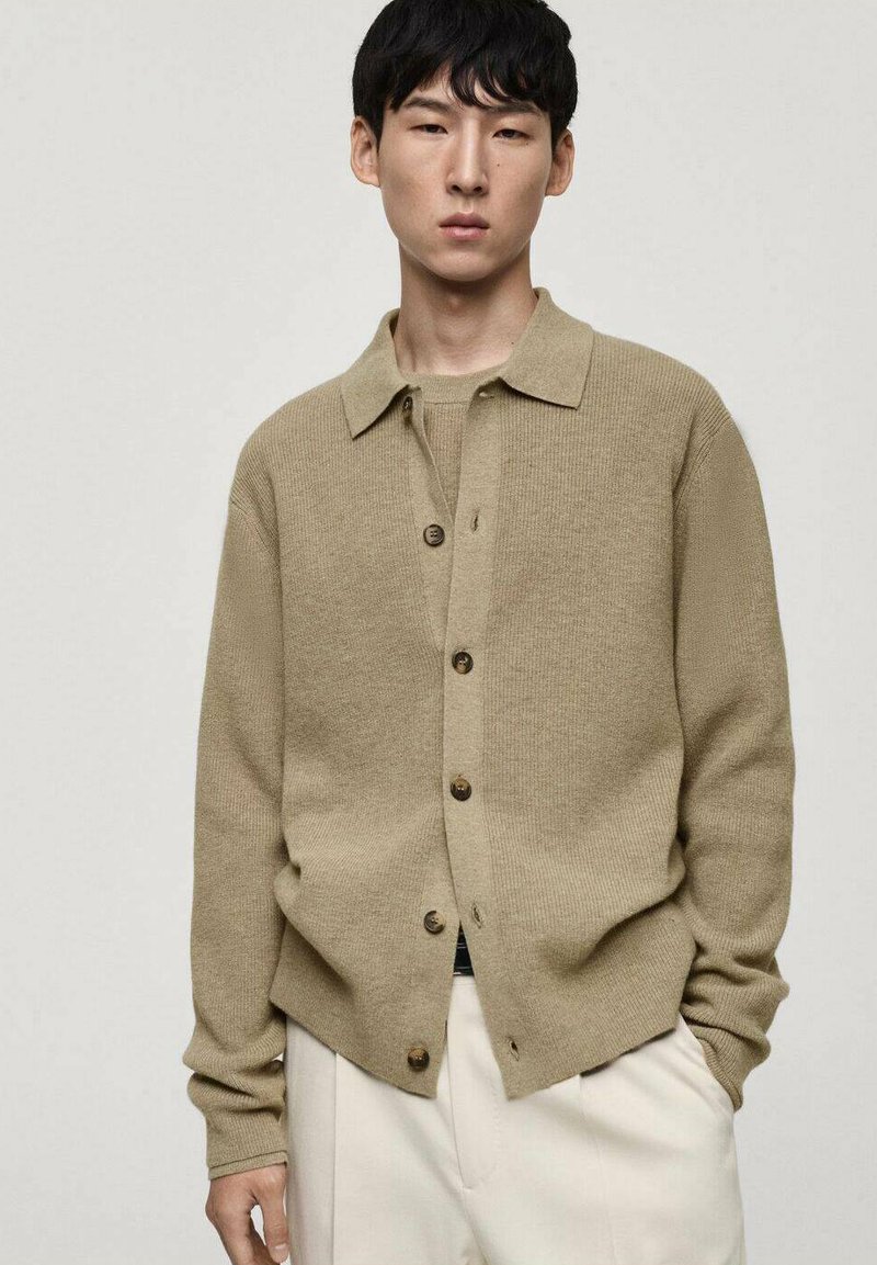 Cardigan lavorato a maglia beige con chiusura a bottoni, colletto aperto e maniche lunghe, abbinato a pantaloni crema, creando un look a strati.