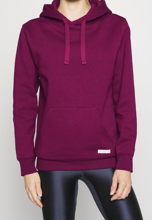 Person wearing a plain dark purple hooded sweatshirt with front pockets and black shiny leggings, shown from neck to hips.