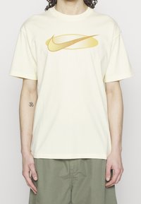 Camiseta de algodón crema con un gráfico del logo de Nike en amarillo y detalle punteado, ajuste relajado y mangas cortas. Combinada con pantalones cortos de color verde oliva.
