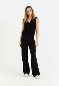 Zwarte gebreide mouwloze top met V-hals en knopen, gecombineerd met zwarte wijde broek. Model draagt witte sneakers, tegen een effen achtergrond.