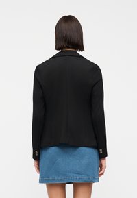 Femme aux cheveux bruns raides mi-longs, portant un blazer noir ajusté et une jupe en jean bleu, vue de dos sur un fond uni.