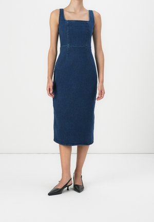 Kobieta nosząca dopasowaną, bezrękawnikową ciemnoniebieską sukienkę midi z denimu z kwadratowym dekoltem i czarnymi, spiczastymi sandałami na obcasie typu slingback.