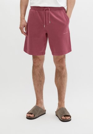 Mężczyzna w bordowych szortach z regulacją w talii i szarych klapkach z odkrytymi palcami, stojący na tle jednolitej, białej ściany.