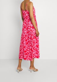 Robe midi fleurie dans des tons roses avec fines bretelles, taille froncée et jupe fluide. Portée avec des talons argentés, présentant une texture de tissu lisse.