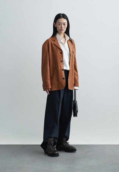 Blazer in suede marrone con grandi tasche, camicia bianca sotto, pantaloni navy a gamba larga e stivali neri robusti. Borsa nera con tracolla a catena.