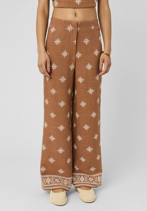 Pantalon marron à jambes larges avec imprimés géométriques blancs et motifs en bordure, porté avec des chaussures beige à enfiler et un top marron assorti.