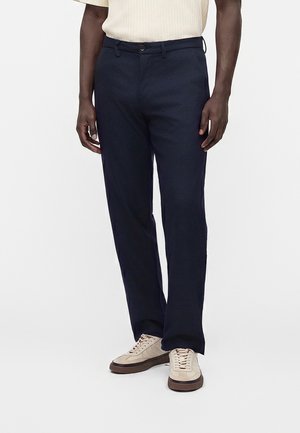 Homme portant un pantalon droit bleu marine foncé avec des baskets beige et une chemise légère à manches courtes, debout devant un fond uni.