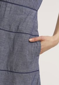 Vestido de lino azul que presenta una superficie texturada, patrones de costura horizontales y un bolsillo lateral. Mano colocada sobre el bolsillo.