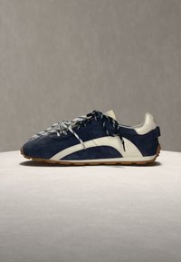 Baskets bleu foncé et blanches à lacets avec une semelle en gomme posées sur une surface claire sur fond gris.