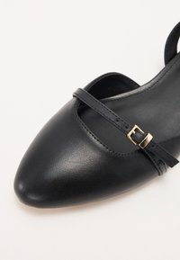 Chaussure en cuir noir avec un bout pointu, dotée d'une fine bride avec une boucle dorée. Texture lisse et design minimaliste.
