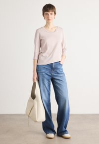 Lichtroze top met driekwart mouwen, gecombineerd met losse blauwe jeans. Model houdt een crèmekleurige schoudertas vast en staat op een lichte, gestructureerde ondergrond.