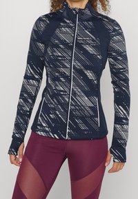 Marinblå idrottsjacka med ett geometriskt grått mönster, hög krage, framdragkedja och tummhål; matchad med bordeaux leggings med meshdetaljer.