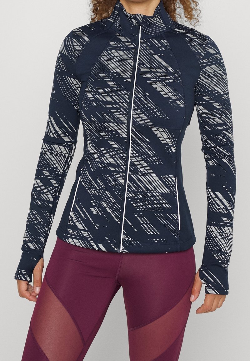 Marinblå idrottsjacka med ett geometriskt grått mönster, hög krage, framdragkedja och tummhål; matchad med bordeaux leggings med meshdetaljer.