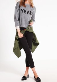 Šedá mikina s nápisem "YEAH" z černých flitrů, černé úzké kalhoty a černé loafers. Olivově зелена bunda uvázaná v pase.