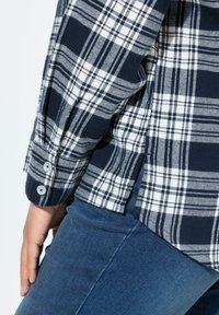 Nahaufnahme einer Person, die ein kariertes Hemd in Marineblau und Weiß mit geknöpften Manschetten und blauen Jeans trägt, die Hand an der Seite ruhend.
