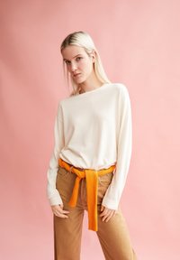 Cream long-sleeve top paired with tan wide-leg pants, accentuated by a textured orange belt. The background is pink.