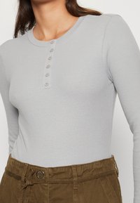 Maglietta a maniche lunghe grigio chiaro a coste con scollatura rotonda e sette bottoni sul davanti. Indossata con pantaloni marroni a vita alta.