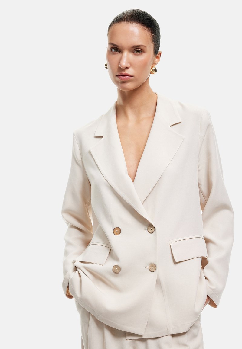 Blazer croisé en tissu beige clair avec des revers, deux boutons en bois et des poches latérales, présentant une coupe décontractée.