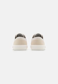 Zapatillas de lona blancas con acentos de tela beige en el talón. Suelas de goma gruesas y detalles cosidos alrededor del collar y los laterales.
