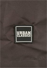 Fekete téglalap alakú folt, amelyen "URBAN CLASSICS" felirat olvasható vastag fehér betűkkel, egy textúrált barna szövet háttérre varrva.