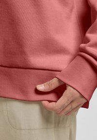 Felpa rosa con una finitura testurizzata, caratterizzata da orli e polsini a costine. Una mano sta sistemando l'orlo, rivelando uno sfondo beige.