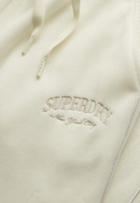 Kremowy hoodie o miękkiej, prążkowanej teksturze, z haftowanym logo "SUPERDRY" w jasnoszarym kolorze nad napisem "New York". Dostosowywane sznurki.