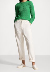 Kvinna som bär en grön stickad tröja, vita byxor med hög midja och framfickor, uppvikta ärmar och vita snöriga sneakers.