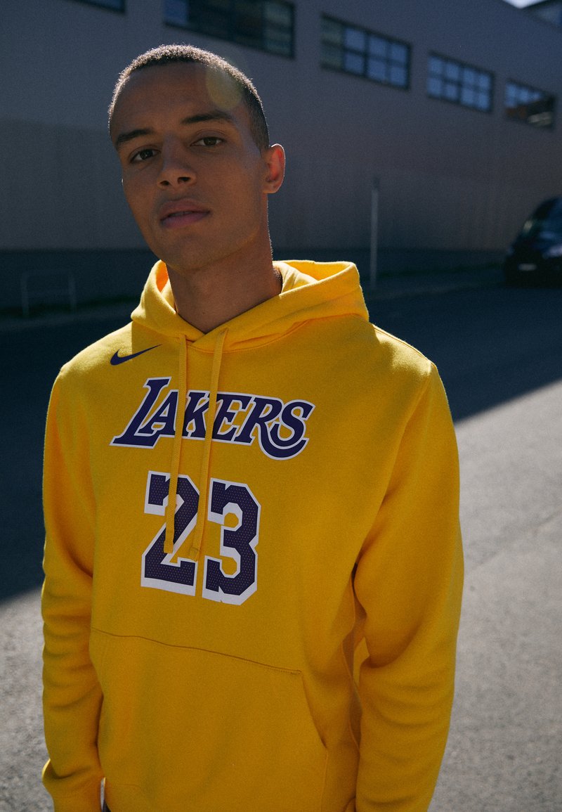 Sweatshirt amarelo com "LAKERS" e "23" impressos em roxo. O tecido é macio, com um bolso frontal e capuz com cordão. Design simples.